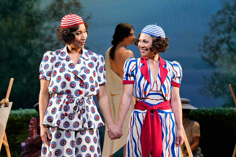 Two women dressed in 1920s costumes stand side by side holding hands and looking at each other. Stage image from "Cosi Fan Tutte" opera
