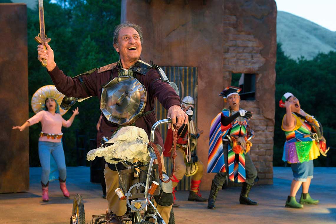a man stands in the foreground with a motley outfit on smiling widely. scene from :quixote nuevo"