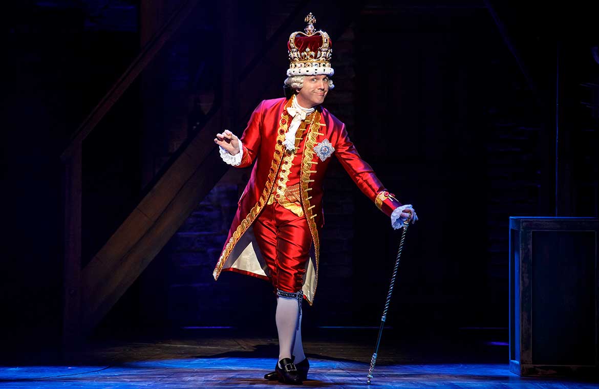 King George from the production of Hamilton stands on stage in a red outfit from the 1700s and a crown