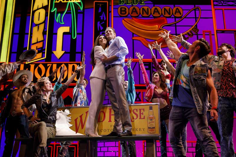 Image from pretty woman the musical. Vivian and Edward stand in the center standing on a park bench holding each other with other cast members surrounding them