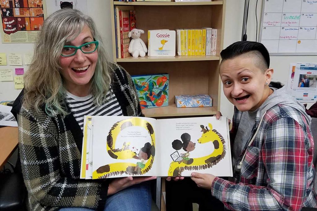 Two women sit together smiling and holding a children's book