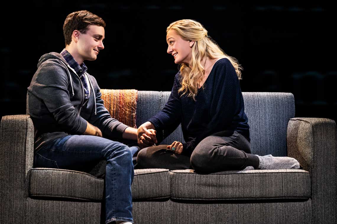 on stage image of dear evan hansen. a young man sits on a couch talking to a young woman.