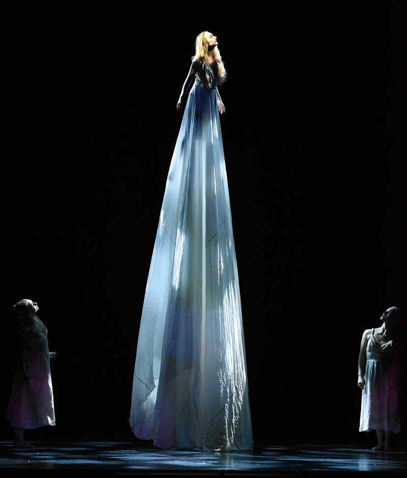 a woman on stilts wears a long white dress while two other woman look up at her from below
