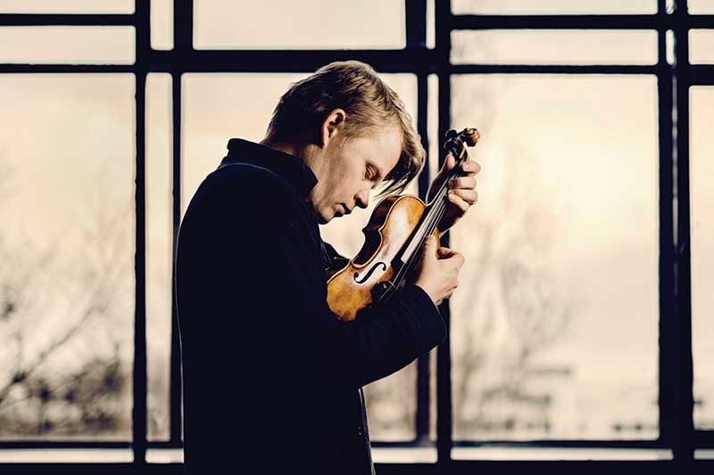 the artist pekka kuusisto stands in front of windows bowing his head over his violin