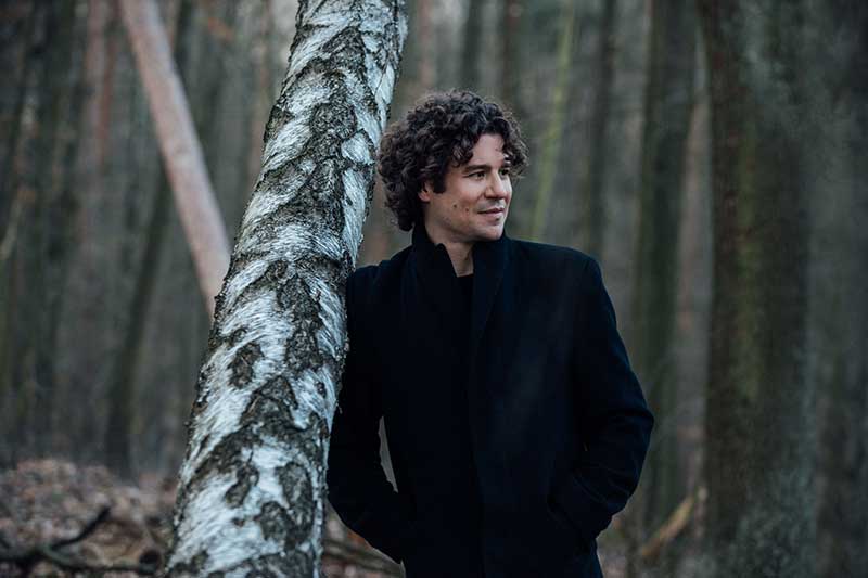 conductor robin ticciati stands in the woods against a tree