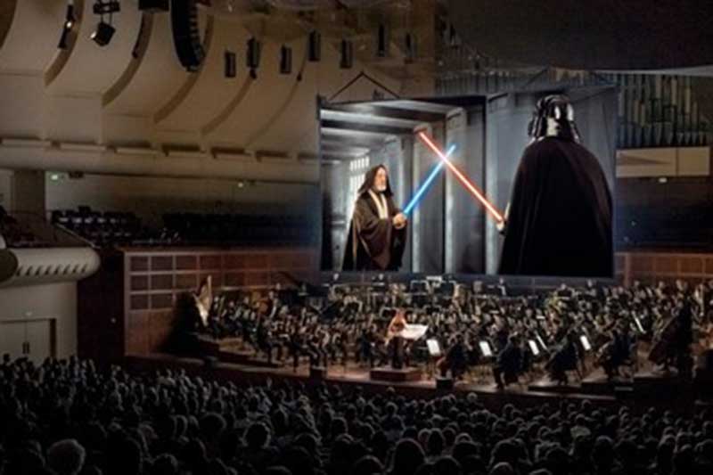 an audience sits and watches star wars on a screen with an orchestra playing below