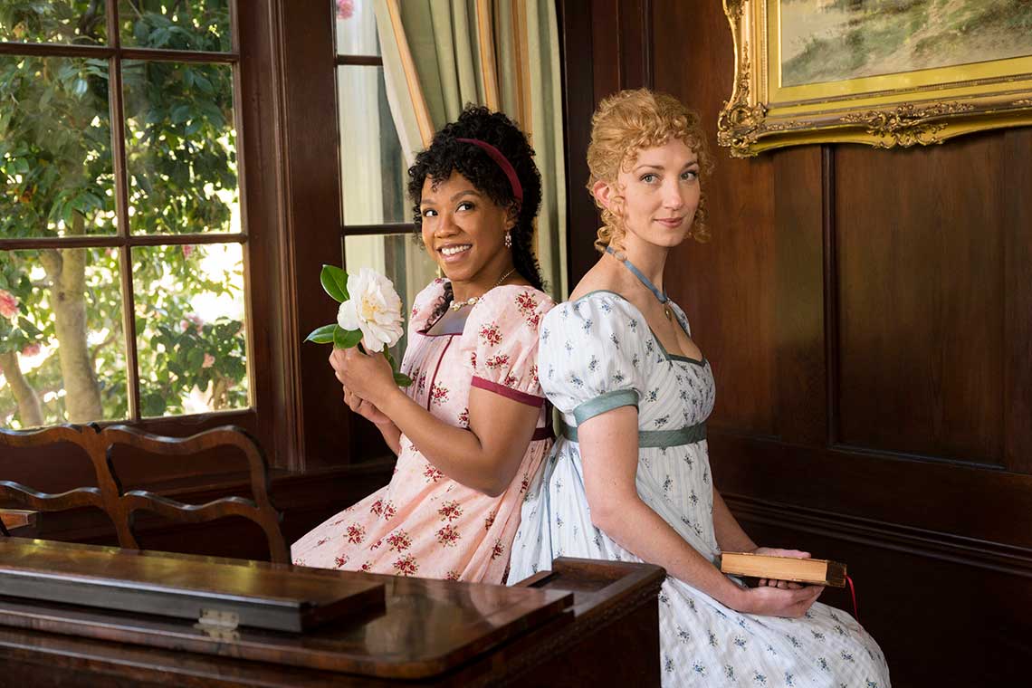 two young women sit back to back on a piano bench in regency era dresses.