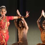 a woman in the forefront wears traditional south indian garb while two dancers behind her kneel with their arms crossed in the air