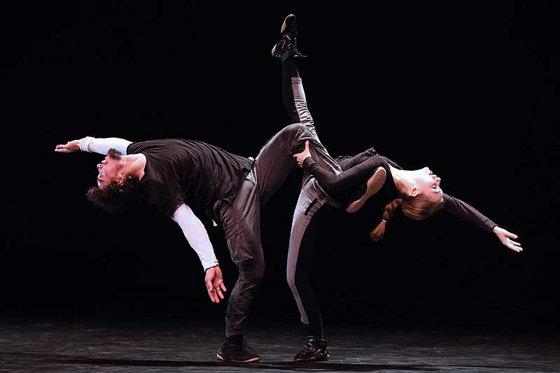 one male dancer and one female dancer face each other with one leg lifted in the air and their backs bent