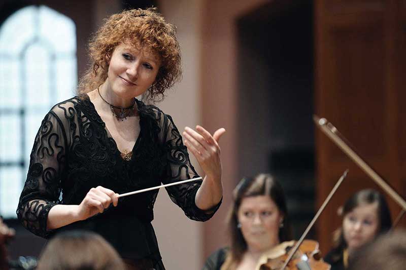 conductor Jeanette Sorrell stands and smiles conducting musicians