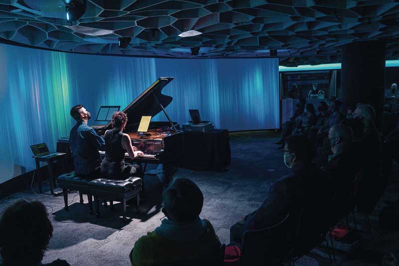 in the octave 9 space, two musicians sit playing piano together in front of a small audience