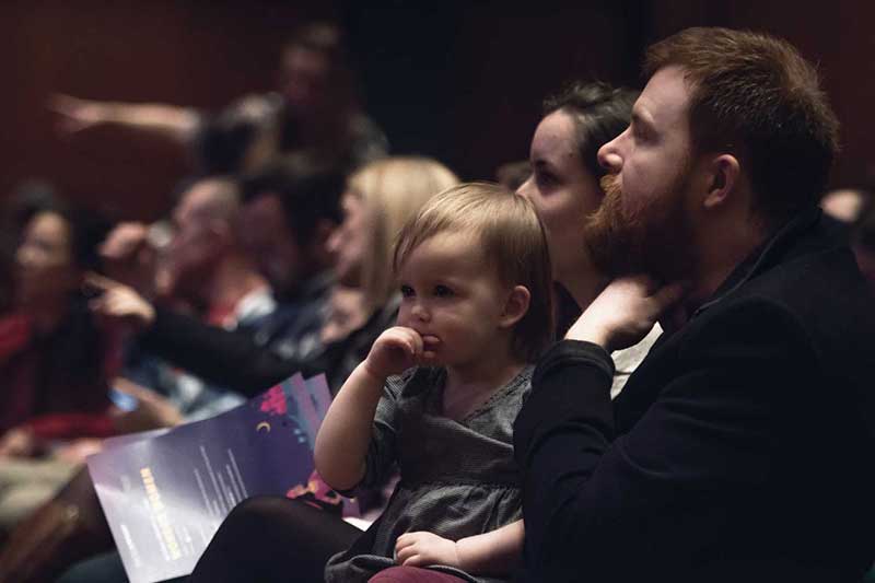 a father sits with his young child on his lap in the audience watching performers on stage