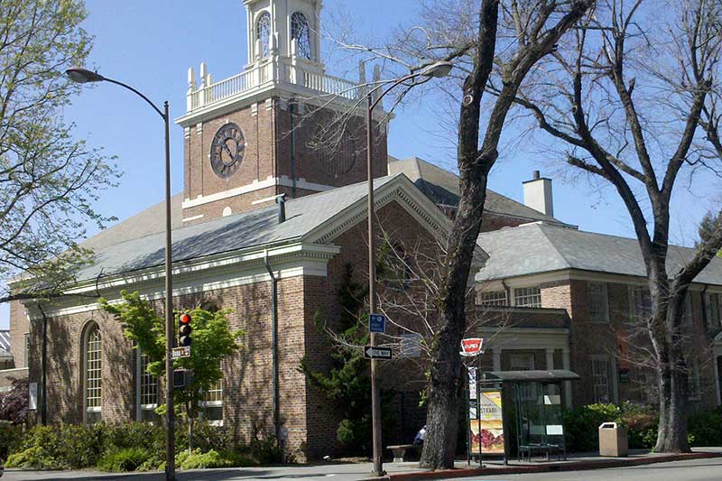 the exterior of a brick church with a clock on the tower and a white bell tower with trees out front
