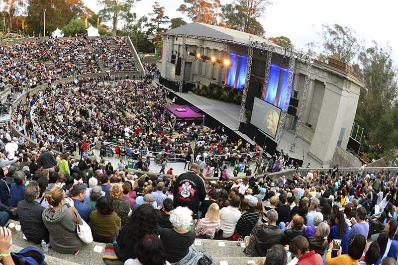 a large audience sits in an outdoor amphitheater watching a performance onstage
