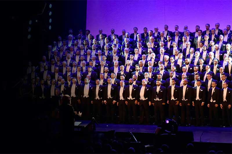a large choir stands singing dressed in tuxedos