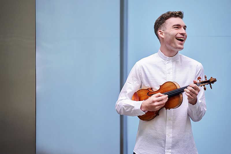 violinist Alexi Kenney wears a whites shirt smiling with Violin