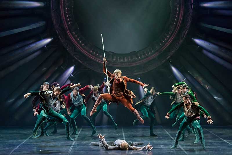 a ballet dancer in the forefront leaps and holds a spear over a dancer on the floor with background dancers behind them