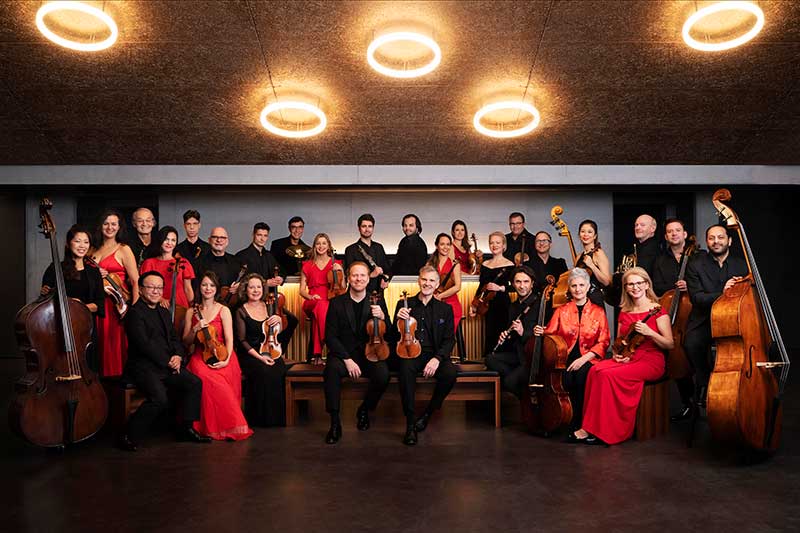 the zurich chamber orchestra stands wearing red and black with their instruments