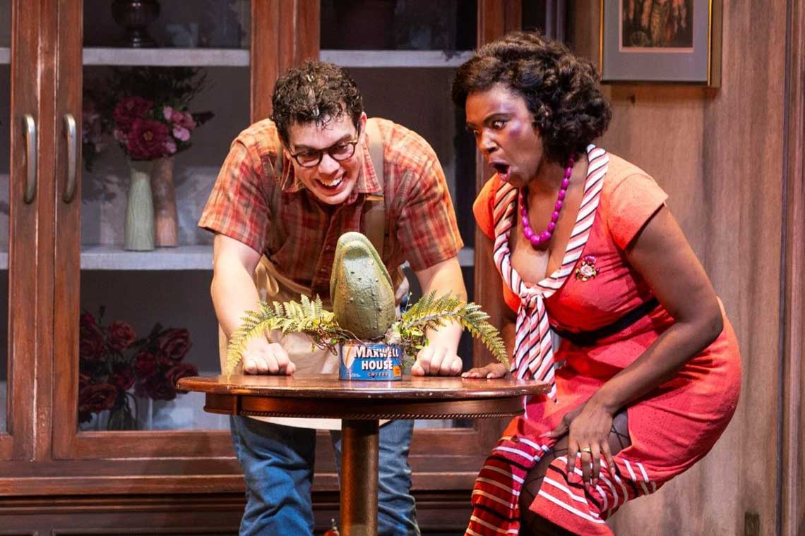 a white man in glasses and a black woman in a pink dress crouch behind a table where a plant sits on top