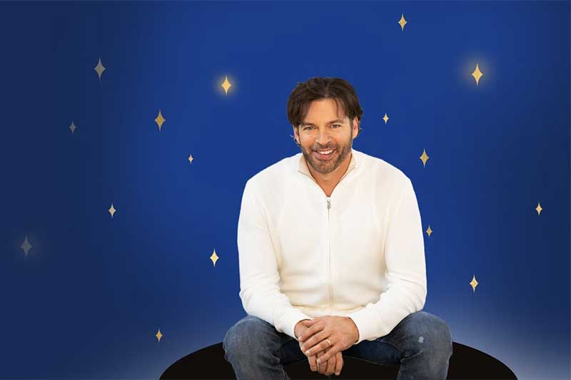 Harry Connick Jr sits in a white shirt in front of a background of stars