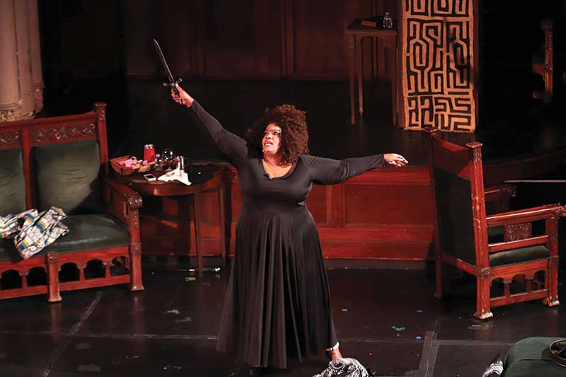 Performer Debra Ann Byrd stands on stage in a black dress with her arms raised above her head with a dagger in one hand.