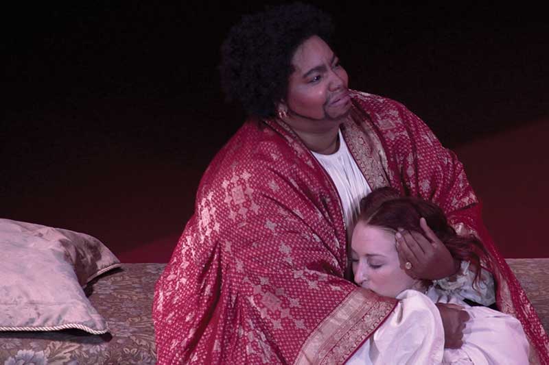 Performer Debra Ann Byrd sits on the ground with a woman's head in her lap wearing a red and gold robe.