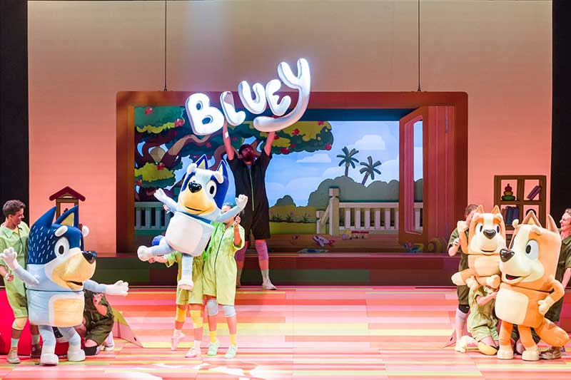 Puppeteers on stage have large dog puppets underneath a Bluey sign.