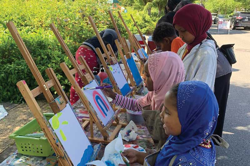three young girls wearing hijabs are outside painting on easels. 