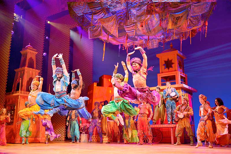 On a very colorful stage, dancers wearing different colors of arabian outfits leap through the air with their hands above their heads. Agraba is in the background.