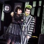 On stage Lydia wears a black lace dress while Beetlejuice in his striped suit wraps his arm around her smiling.