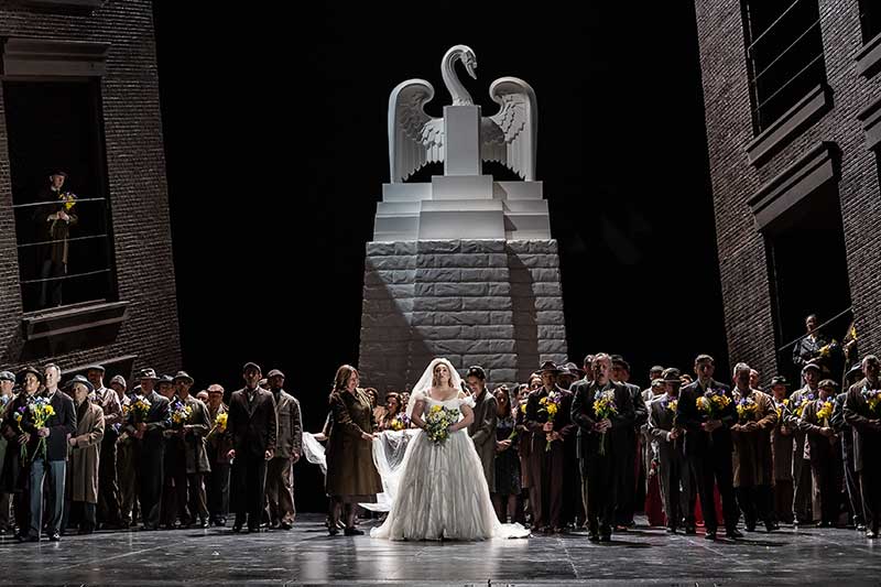 On stage a crowd stands wearing dark clothes in front of a large statue of a swan. In the middle, a woman stands wearing a wedding dress.