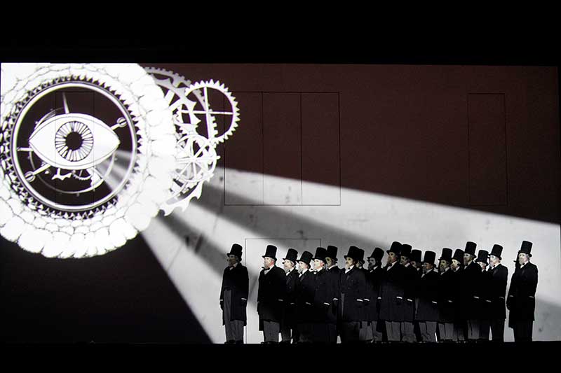 On stage a large group of men in black suits and top hats stand in a line looking at a white eye that hangs above in the background.