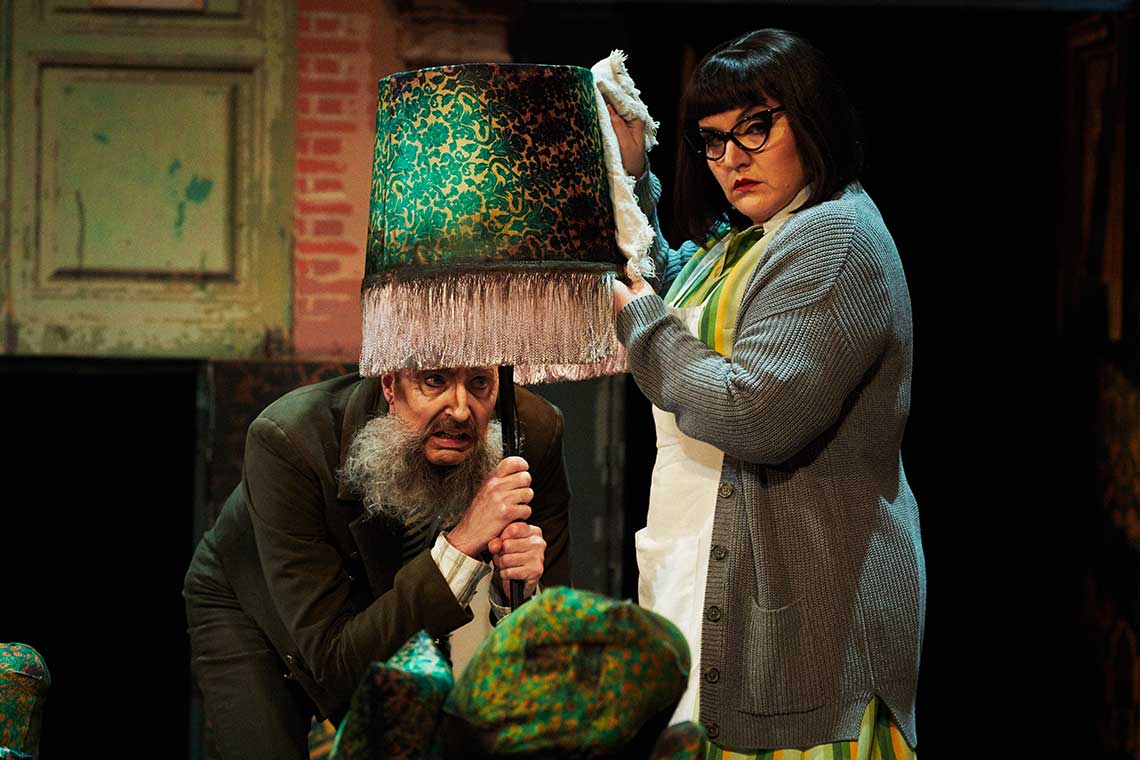 a man with a long beard hunkers down hiding under a lamp while a woman stands above him