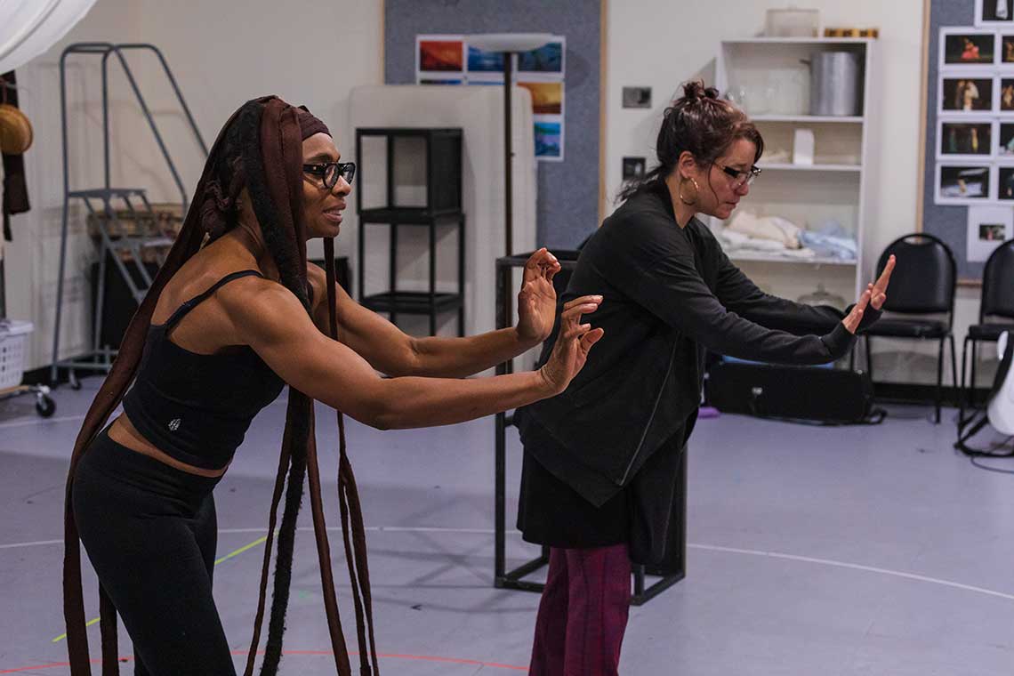 Two actors, a white woman and a black woman, are in rehearsal with their hands extended in front of them as if pushing something.