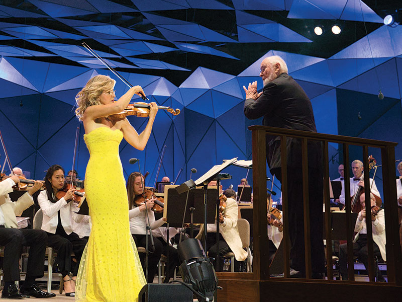 Anne-Sophie Mutter, in a yellow dress, plays violin in front of an orchestra while John Williams conducts