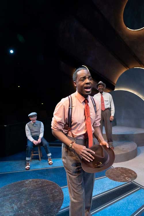 On stage a Black man dressed in a tie and suspenders speaks as he holds his hat.