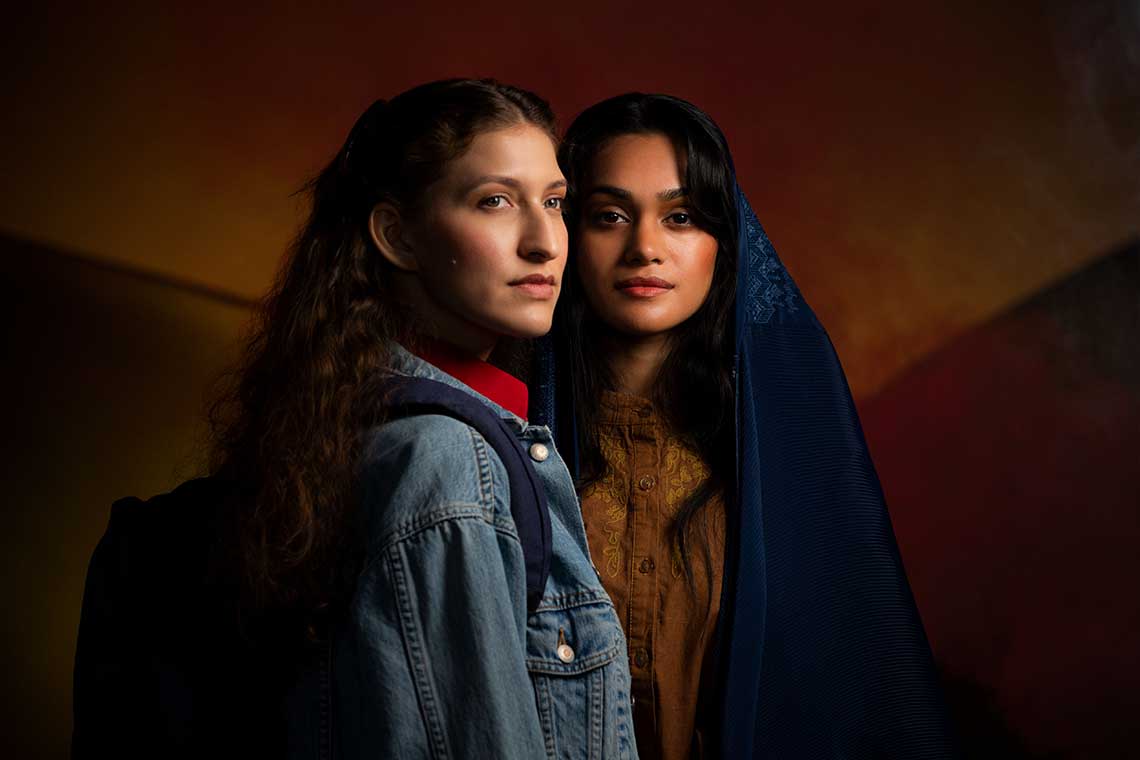 Two women stand side by side looking off into the distance. One wears a jean jacket and the other wears a head scarf.