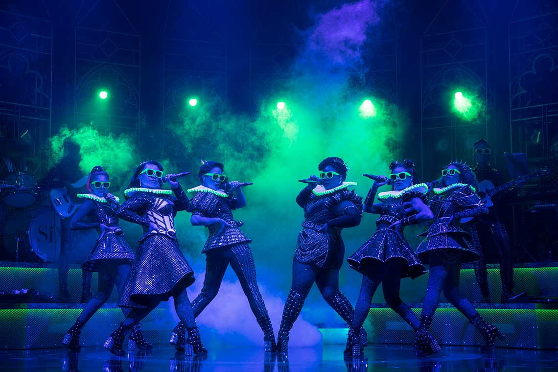 On stage six woman are dressed in modern versions of 16th century dress wearing glowing sunglasses with a smoky green light on them.