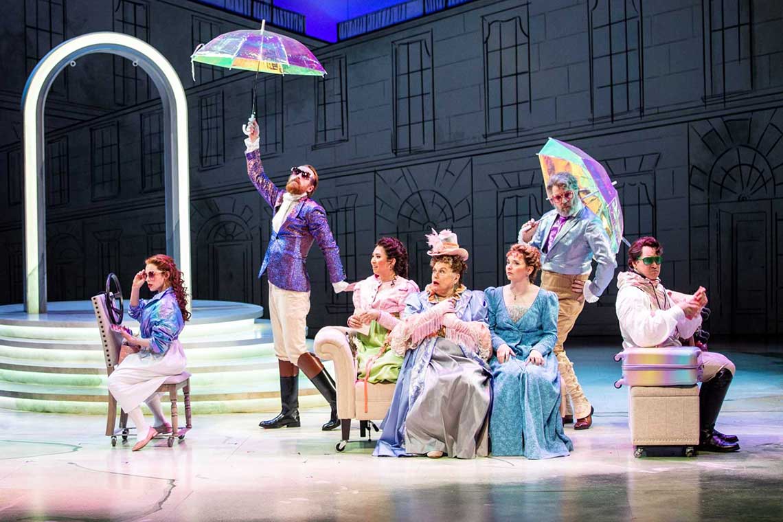 On stage a group of seven men and women sit around wearing sunglasses and 19th century dress with rainbow umbrellas.