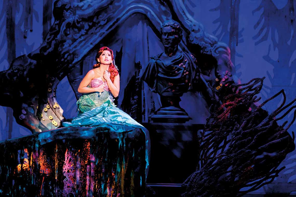 On stage Ariel from the little mermaid sits on a rock underwater singing and looking towards the surface.