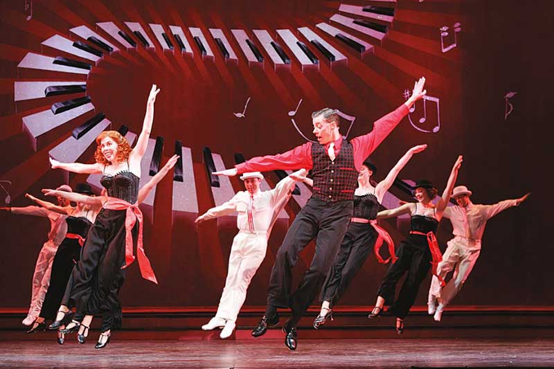 On stage a group of dancers are jumping in air in unison with their arms outstretched. In the background is a piano keyboard.