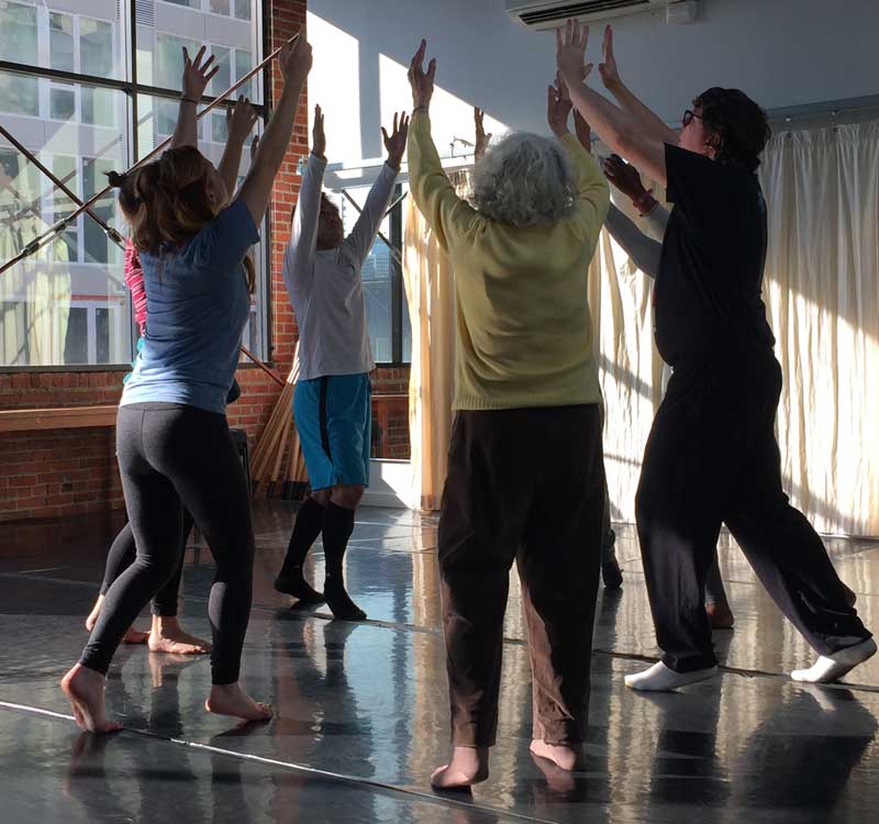 A group of adults stand in a dance studio in a circle with their hands raised above their heads, joining in the middle.