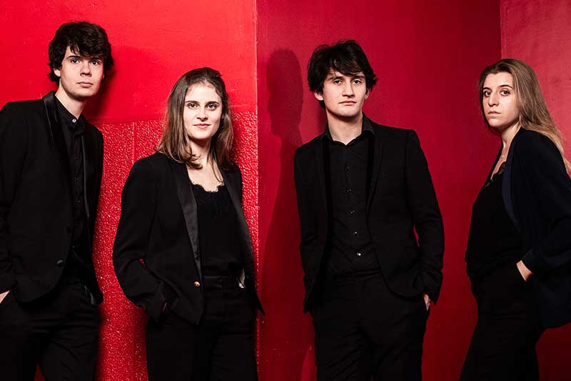 Two young men and two young women wear all black and stand in front of a red background.