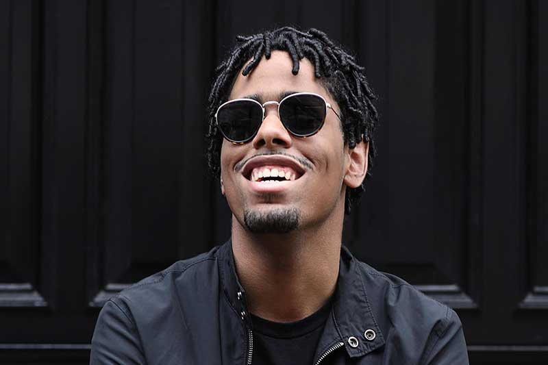 A young black man wears sunglasses and a black coat smiling.
