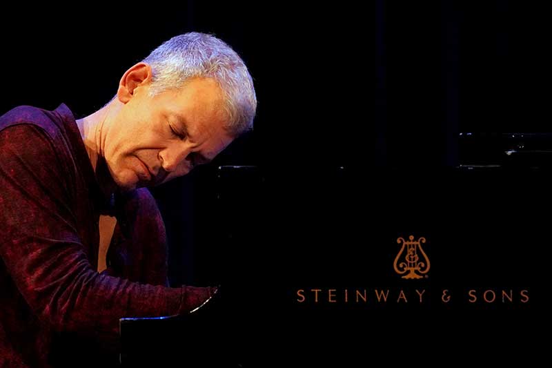 A white man with grey hair sits playing the piano with his eyes closed.