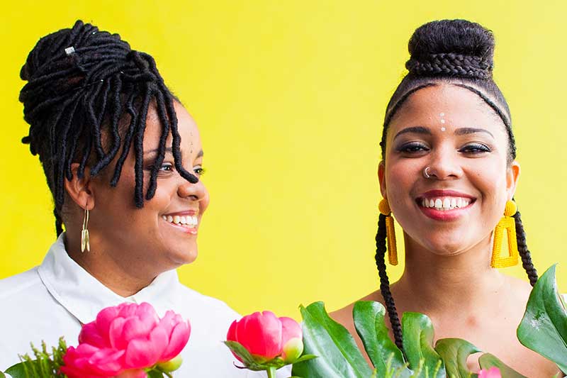 Two black women stand in front of a bright yellow background with just their heads and shoulders showing. Pink flowers rise up next to them.