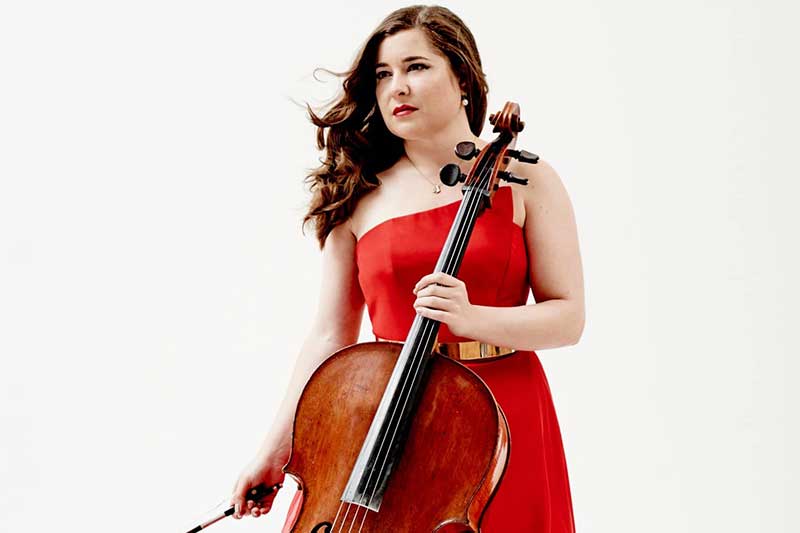 A white woman with brown hair wears a red dress with her cello in front of her.