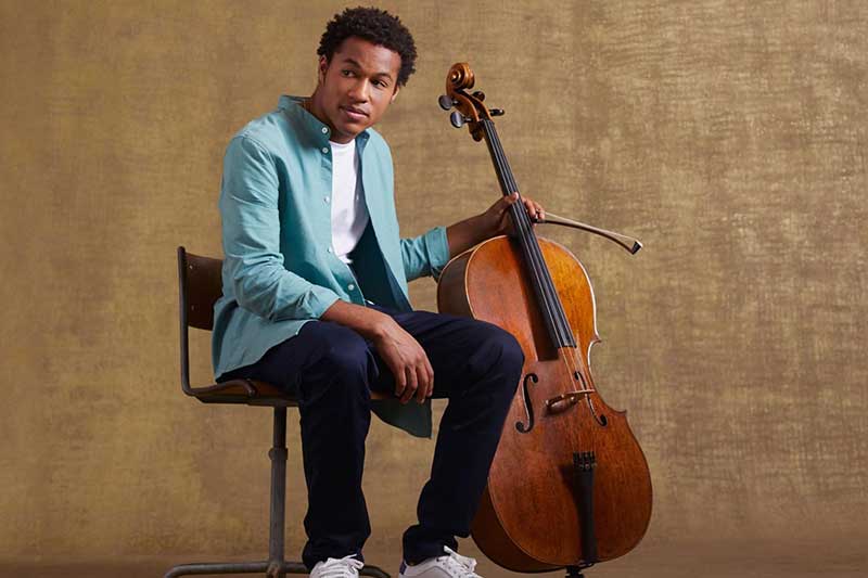 A young black man sits in a chair next to a cello.