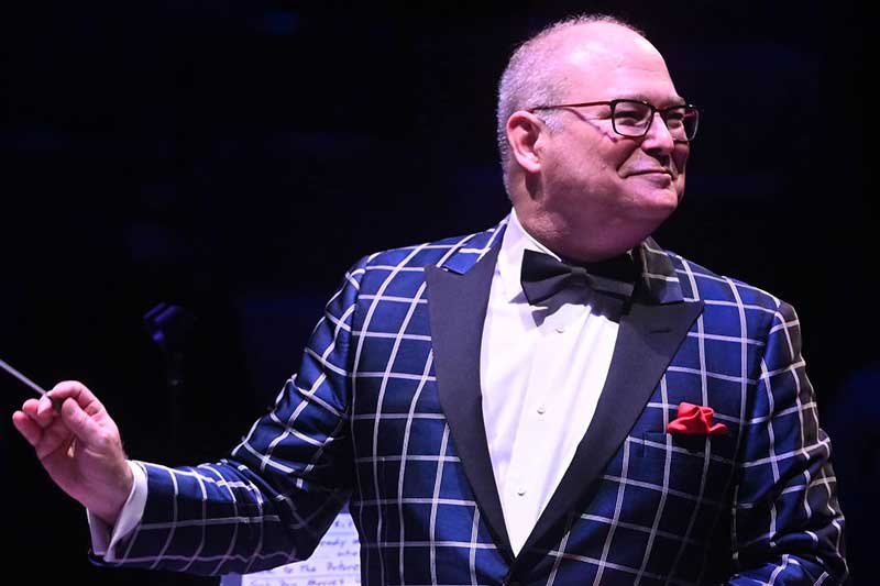 A white man wears a checkered tux smiling as he conducts a symphony.