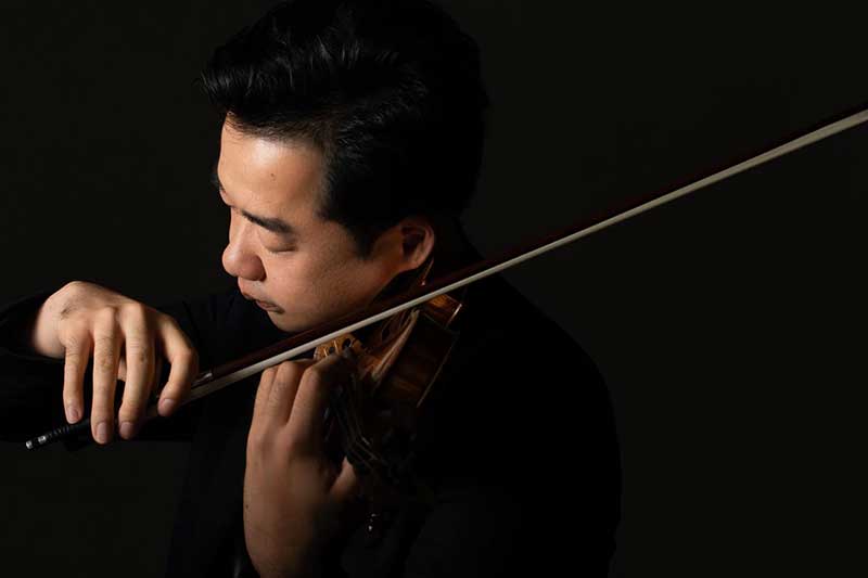 On a black background, an asian man faces the side and holds a violin playing.